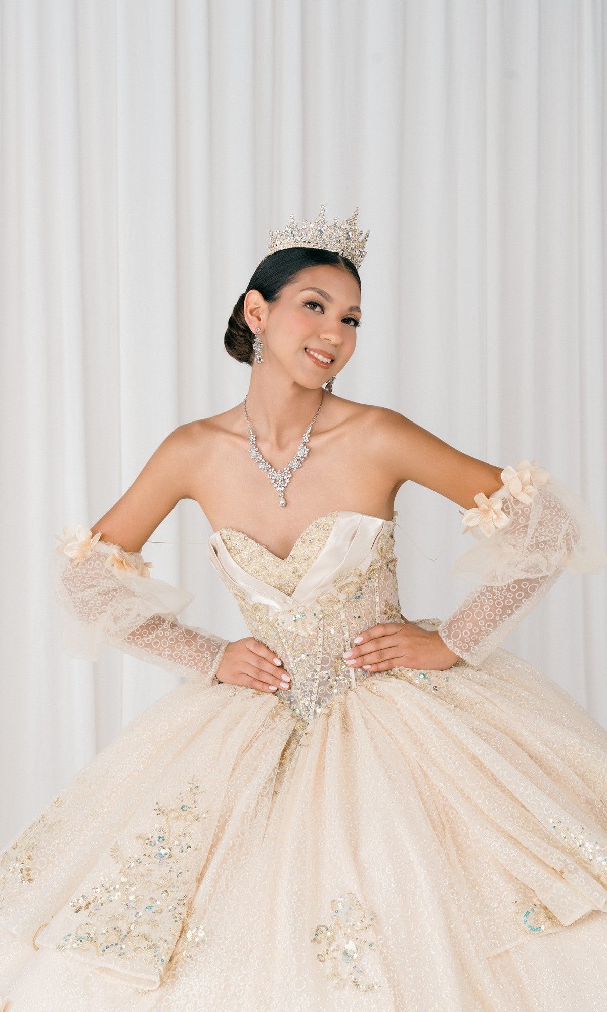 A young woman poses with hands on hips in front of a white curtain, wearing the sparkling cream-colored Quinceanera Dress 8385J By Cinderella Couture with puff sleeves, a tiara, and jewelry.
